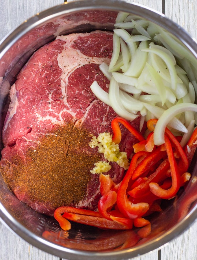 Uncooked Beef roast with chili powder, sliced onions, bell peppers, and garlic inside a pot.