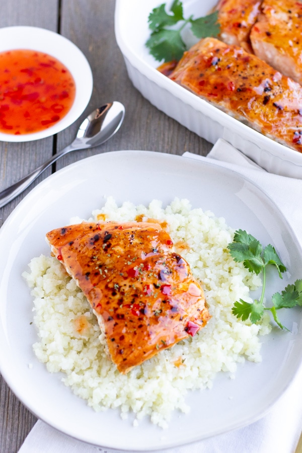 Overhead view of salmon on a plate of cauliflower rice with a side of sweet chili sauce and more salmon in a baking dish.