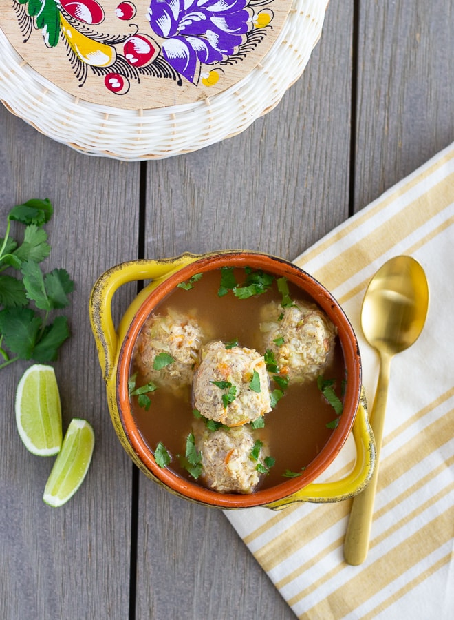 Meatball soup in a yellow bowl with lime wedges and cilantro on the side. Decorative tortilla warmer in the background.