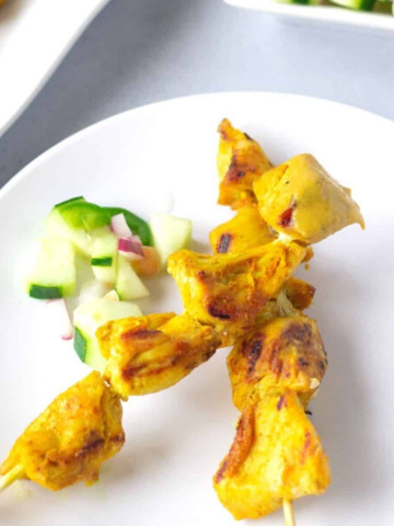 Two chicken skewers on a white plate with some diced cucumbers on the side.