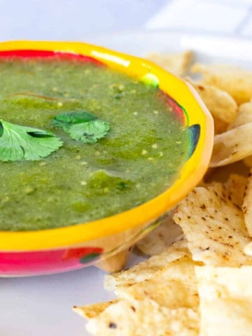 Up close view of salsa verde in a yellow bowl with tortilla chips on the side.