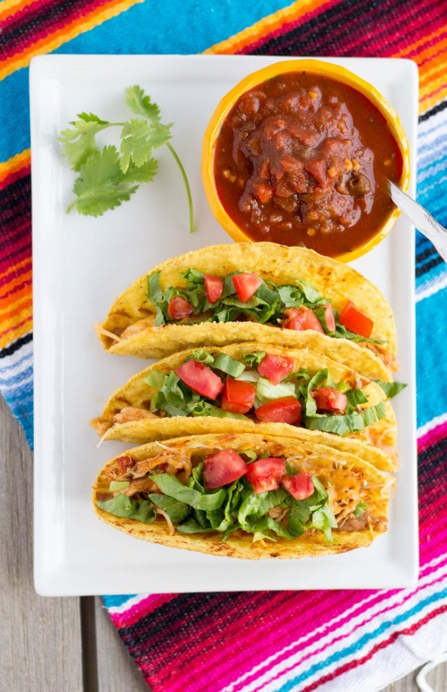 3 tacos on a white plate under a colorful Mexican placemat.