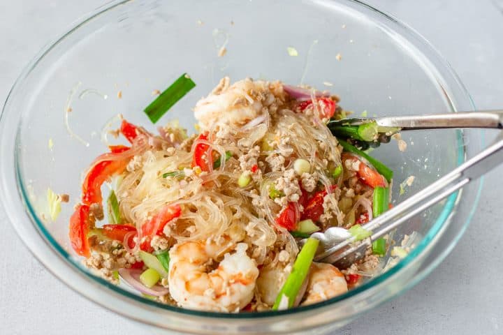 Noodle salad in a glass bowl with tongs.