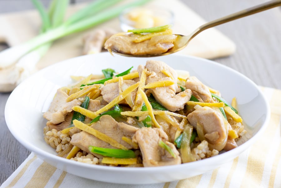 Spoon holding a piece of chicken and ginger over a white bowl.