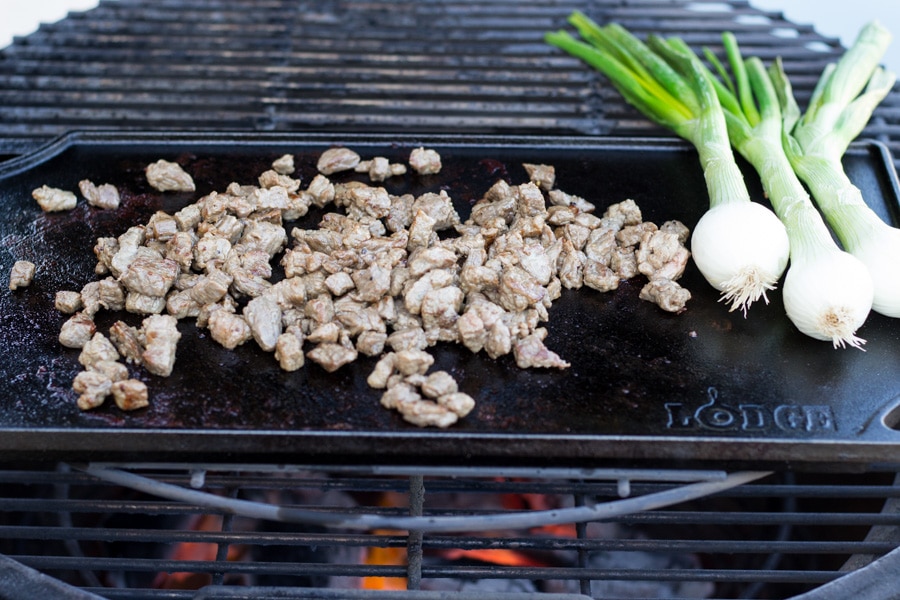 Cast iron flat top on an outdoor grill with steak cooking and onion bulbs.