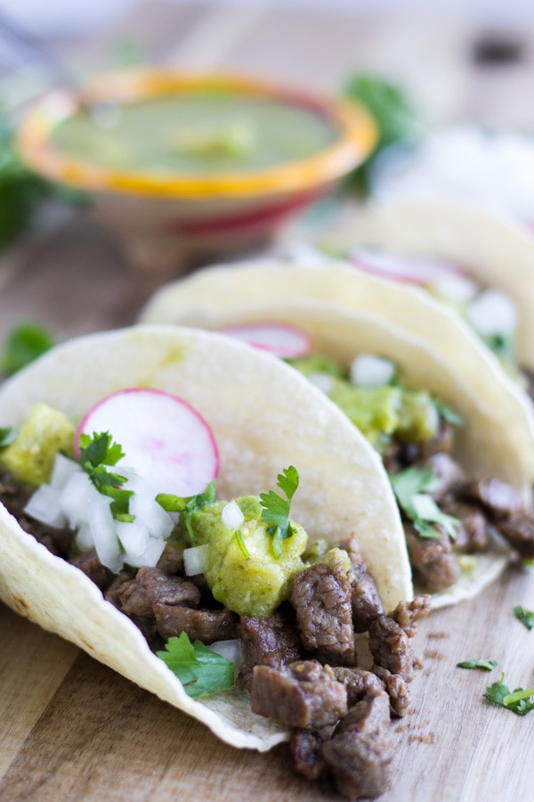 Three Carne Asada Tacos on a cutting board topped with salsa verde, cilantro, onions, and radishes.
