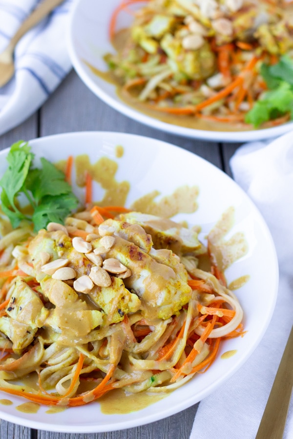 Two white bowls of spiralized zucchini and carrots topped with Thai chicken Satay and a creamy peanut sauce.