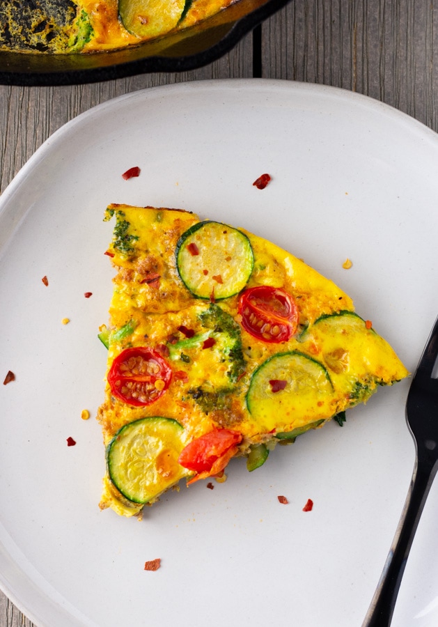 Single slice of frittata on a white plate with a black fork and crushed peppers on top.