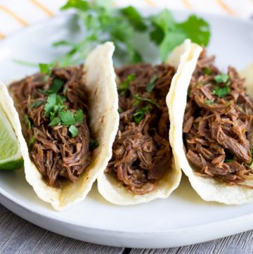 Three shredded beef tacos arranged on a white plate with a slice of lime on the side.