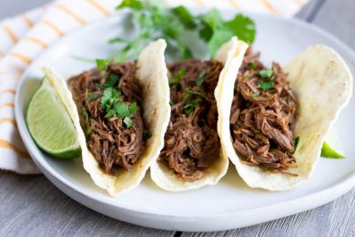 Three shredded beef tacos arranged on a white plate with a slice of lime on the side.