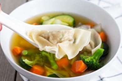 White Soup spoon holding one Wonton above soup with vegetables.