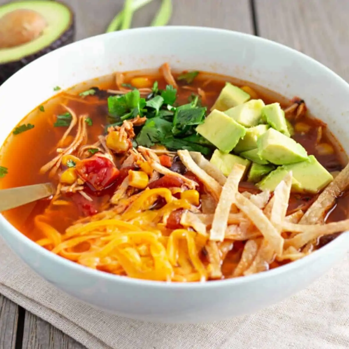Soup in a white bowl topped with cheese, cilantro, avocado, and tortillas strips.