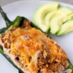 Single stuffed poblano on a white plate with sliced avocado on the side.