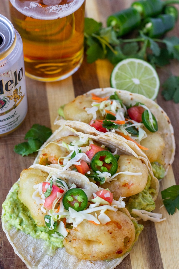 Completed shrimp tacos topped with slaw and jalapenos on a wood board with a glass and can of Modelo on the side.