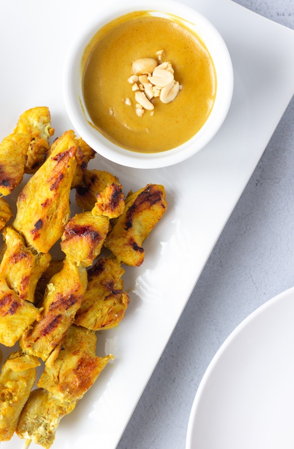 Overhead view of chicken satay skewers on a long plate with a small bowl of peanut sauce.