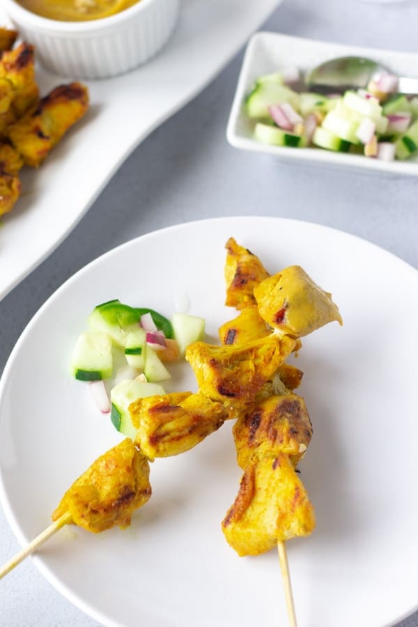 Two skewers of Chicken Satay on a white plate with cucumber relish on the side.