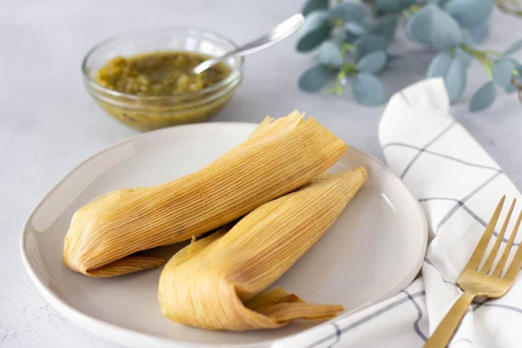 Two tamales wrapped in corn husks on a plate with salsa on the side.