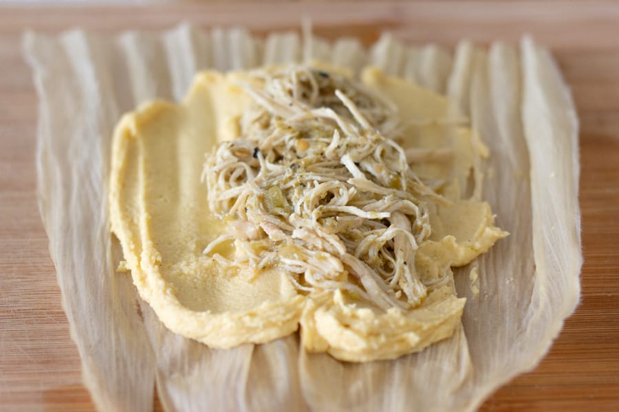 Corn husk with masa and salsa verde chicken on a cutting board.