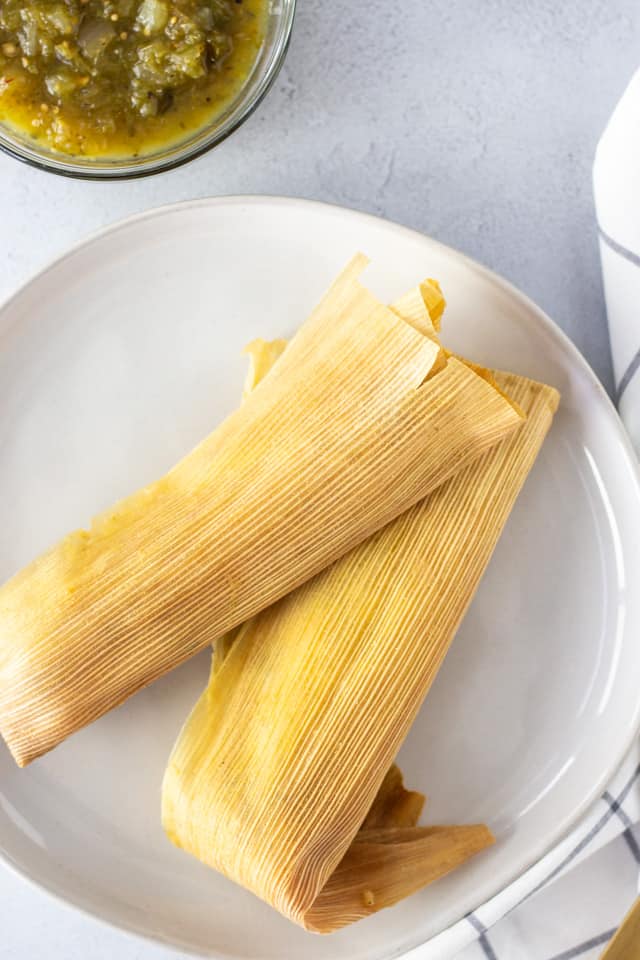 Two tamales wrapped in corn husks on a plate with salsa on the side.