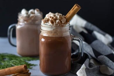 Two clear glasses of Mexican Hot Cocoa, one with a cinnamon stick and the other with marshmallows.