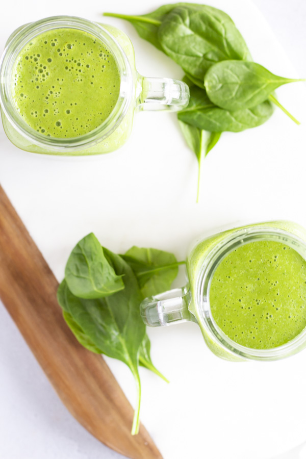 Overhead view of two green smoothies with some spinach on the side.