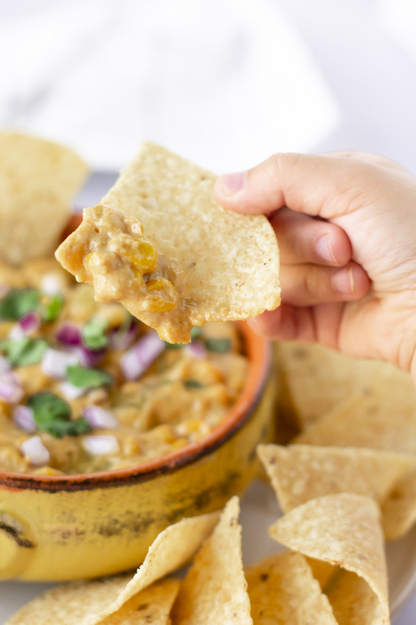 Small hand holding a tortilla chip with some Mexican street corn dip on it.