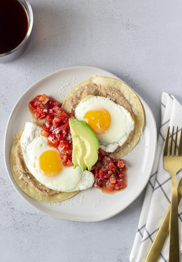 Overhead view of huevos rancheros with a gold fork and knife to the side and a black cup of coffee.