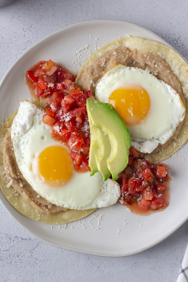 Overhead view of Huevos Rancheros on a white plate and topped with red tomato salsa and avocado
