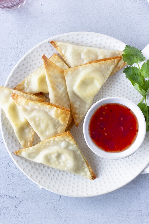 Overhead view of triangle wontons on a plate with a side of sweet chili sauce and garnish of cilantro.