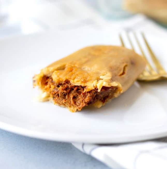 Up close view of a single beef tamale on a white plate with a gold fork on the side.