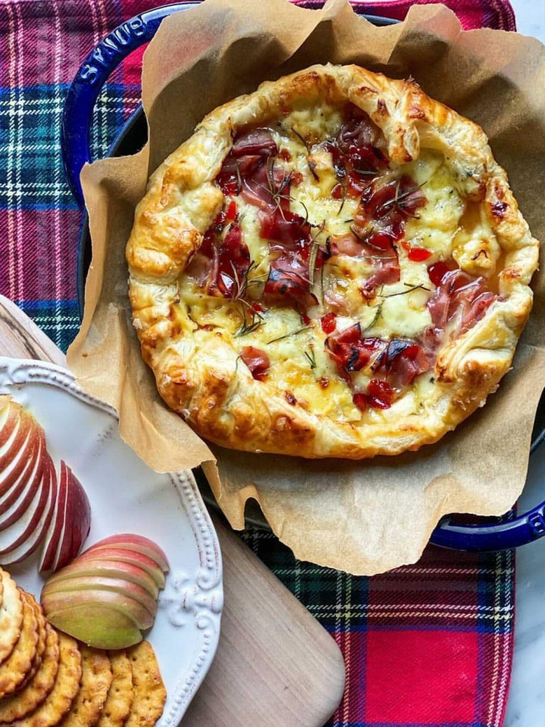 Red plaid napkin with a dish of baked brie inside a cast iron skillet. Apples and Crackers on the side.