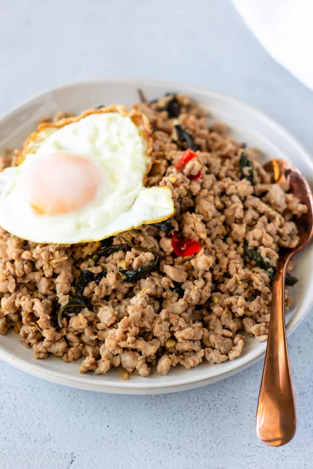Ground pork stir fry on a white plate topped with a fried egg and bronze color spoon on the side.