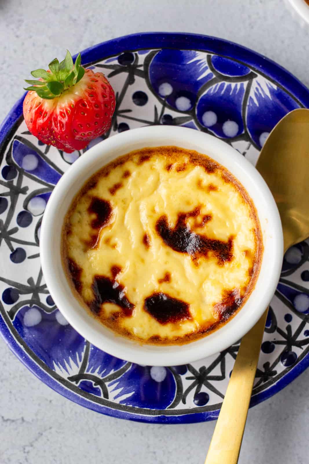 Overhead view of custard in a white ramekin on a blue plate with a gold spoon and strawberry garnish on the side.