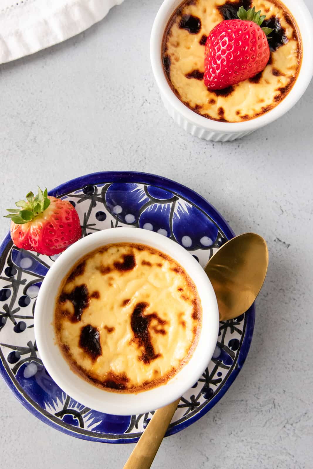 Two white ramekins, one on a blue plate with a gold spoon, and custard garnished with a strawberry.