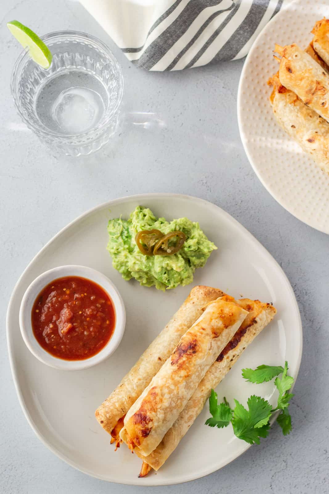 Overhead view of rolled tacos on a plate with a side of salsa and guacamole and glass of water on the side.