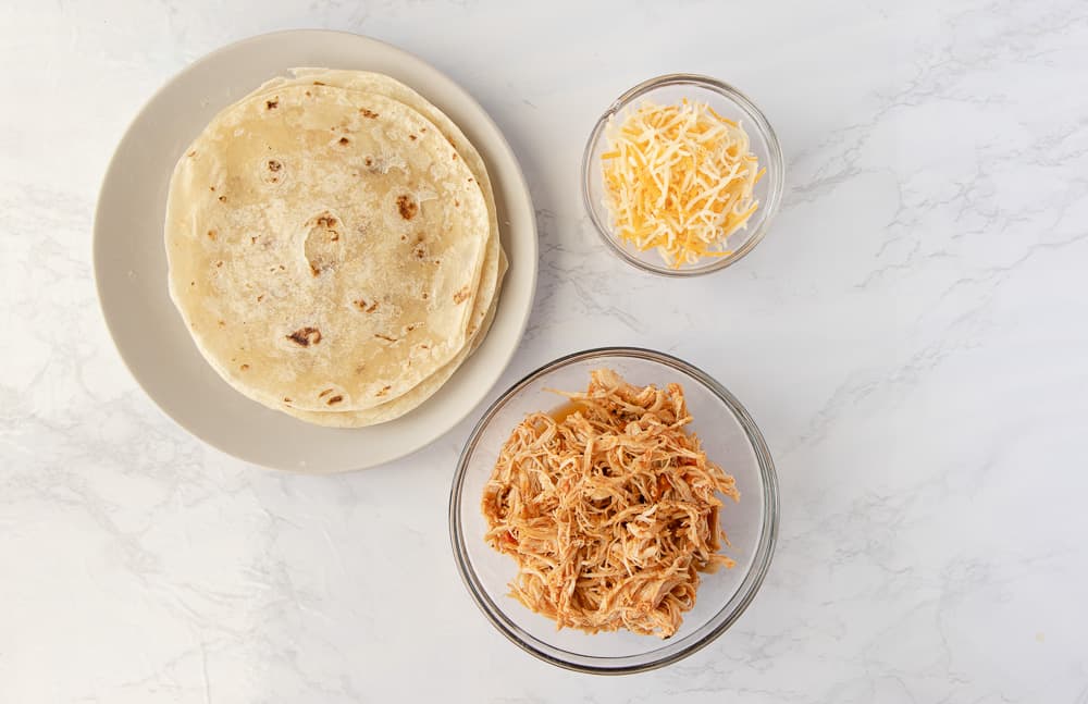 Overhead view of ingredients, flour tortillas, shredded salsa chicken, and mexican shredded cheese.