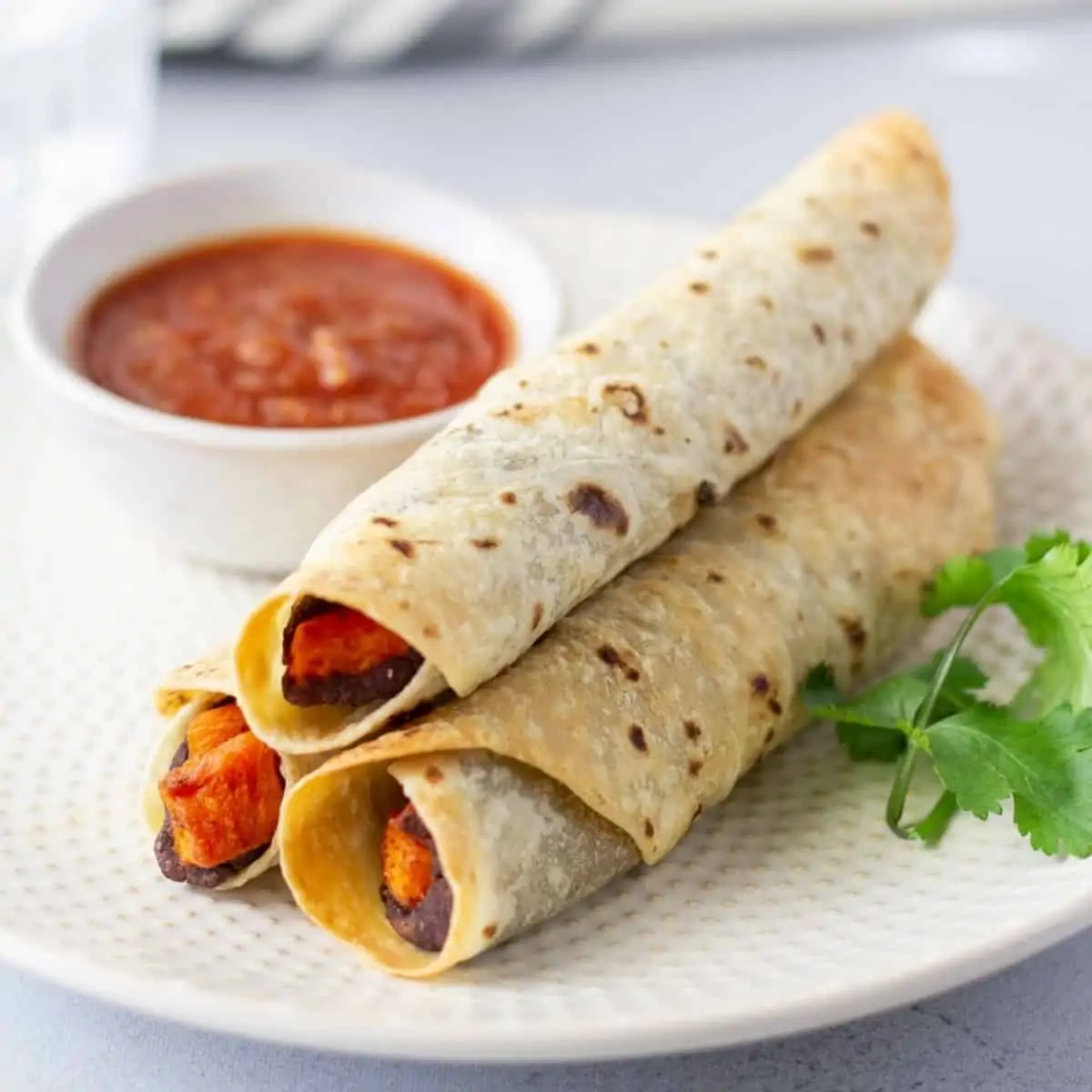 Up close view of 2 flautas on a plate with a side of salsa and a cilantro stem for garnish.
