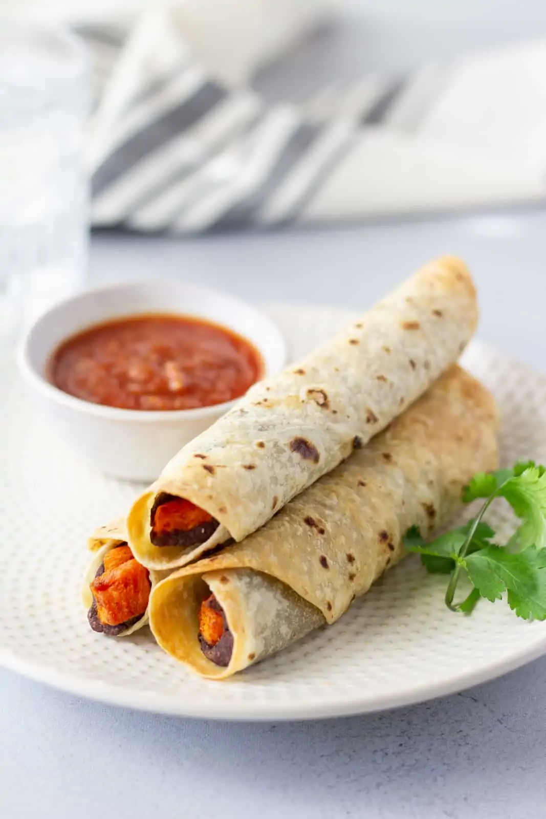 Three rolled tacos on a plate with a side of salsa and garnish of cilantro.