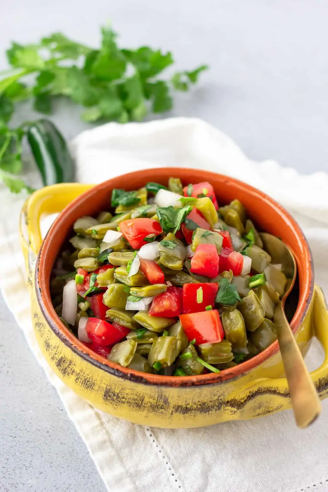 Nopales in a yellow bowl with a gold spoon. Cilantro and jalapeno in the background.