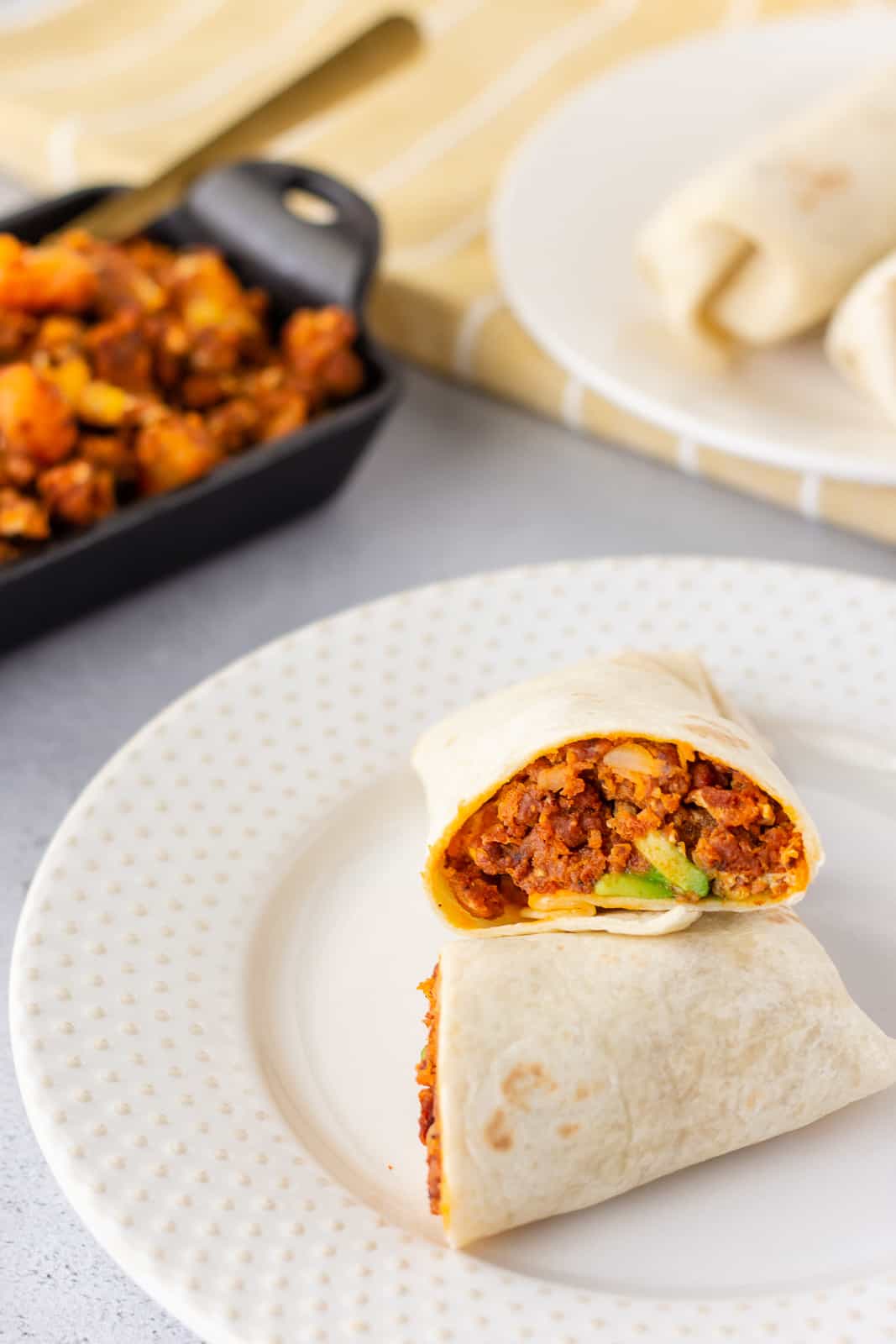 Burrito on plate sliced in half and stacked on each other to show filling. Small skillet in the background with extra filling