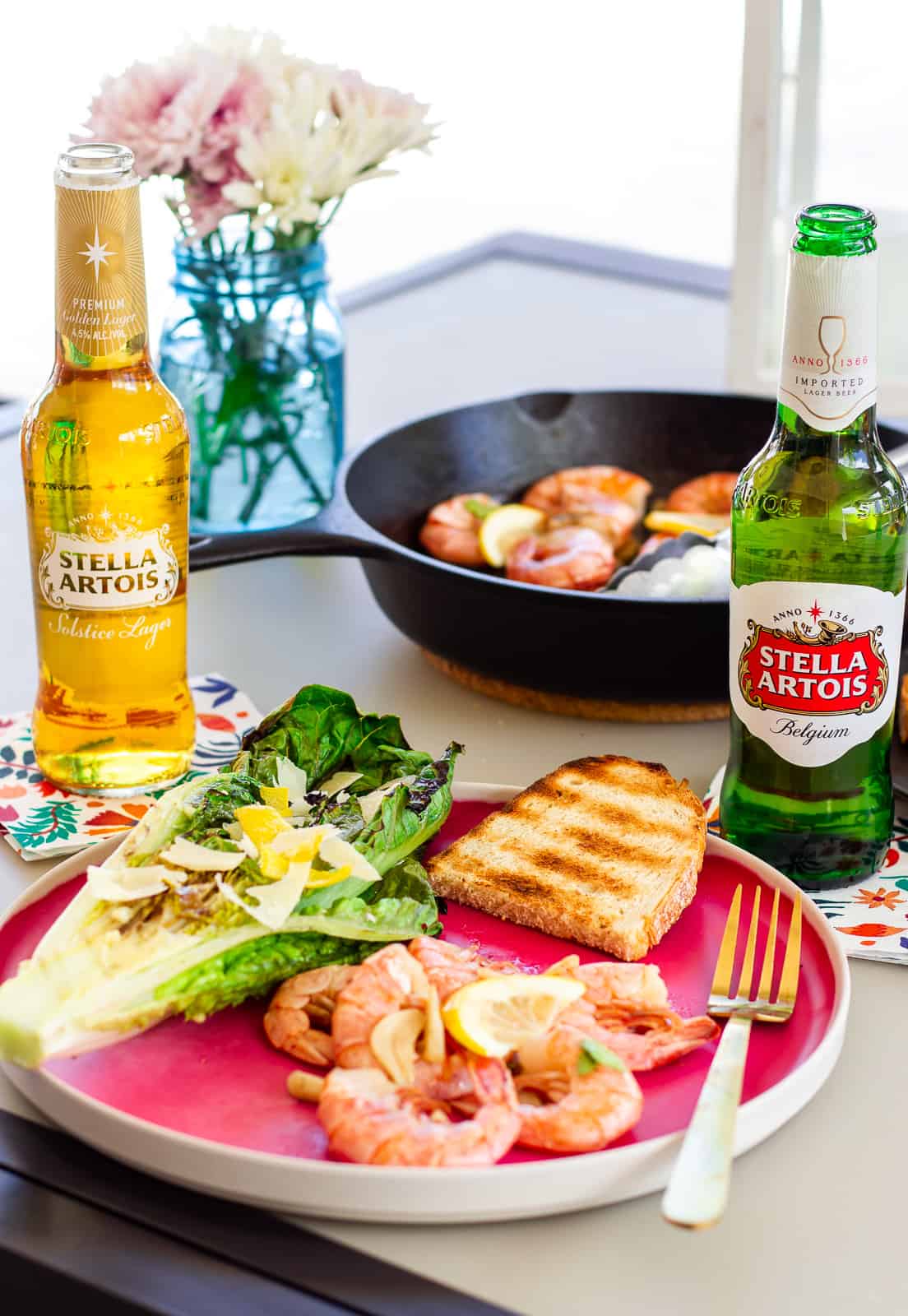 Pink plate with shrimp, slice of grilled bread, halved grilled romaine, and 2 bottles of Stella Artois Beer.