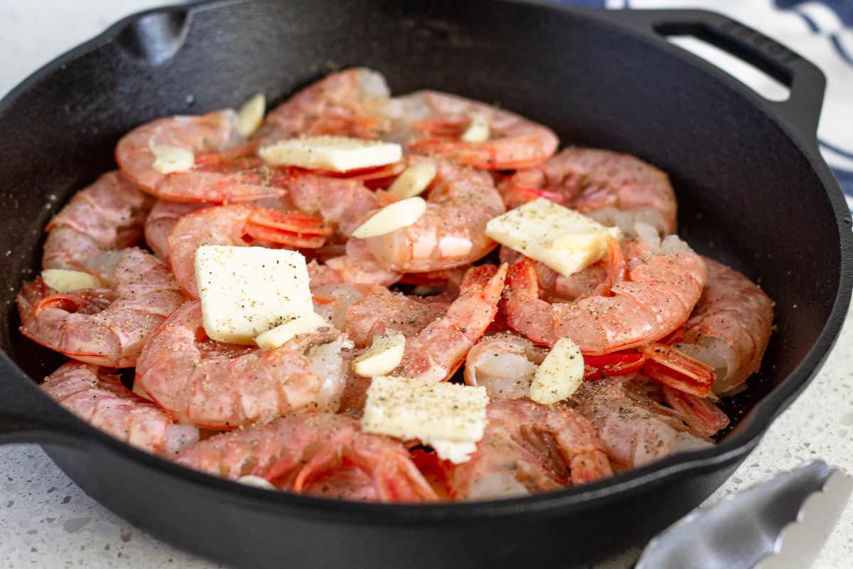 Raw shrimp in a cast iron skillet with slices of butter and sliced garlic on top.