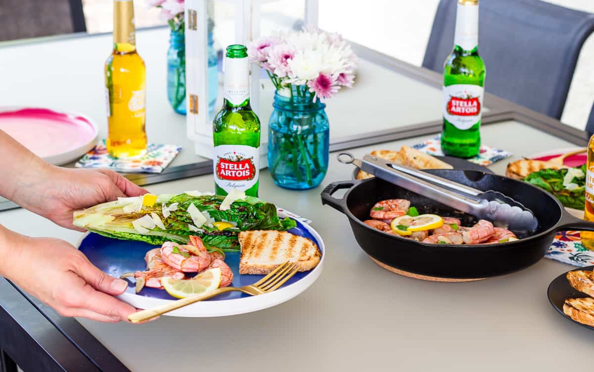 Hand holding a plate above the table with Stella Artois Beers and skillet with shrimp.