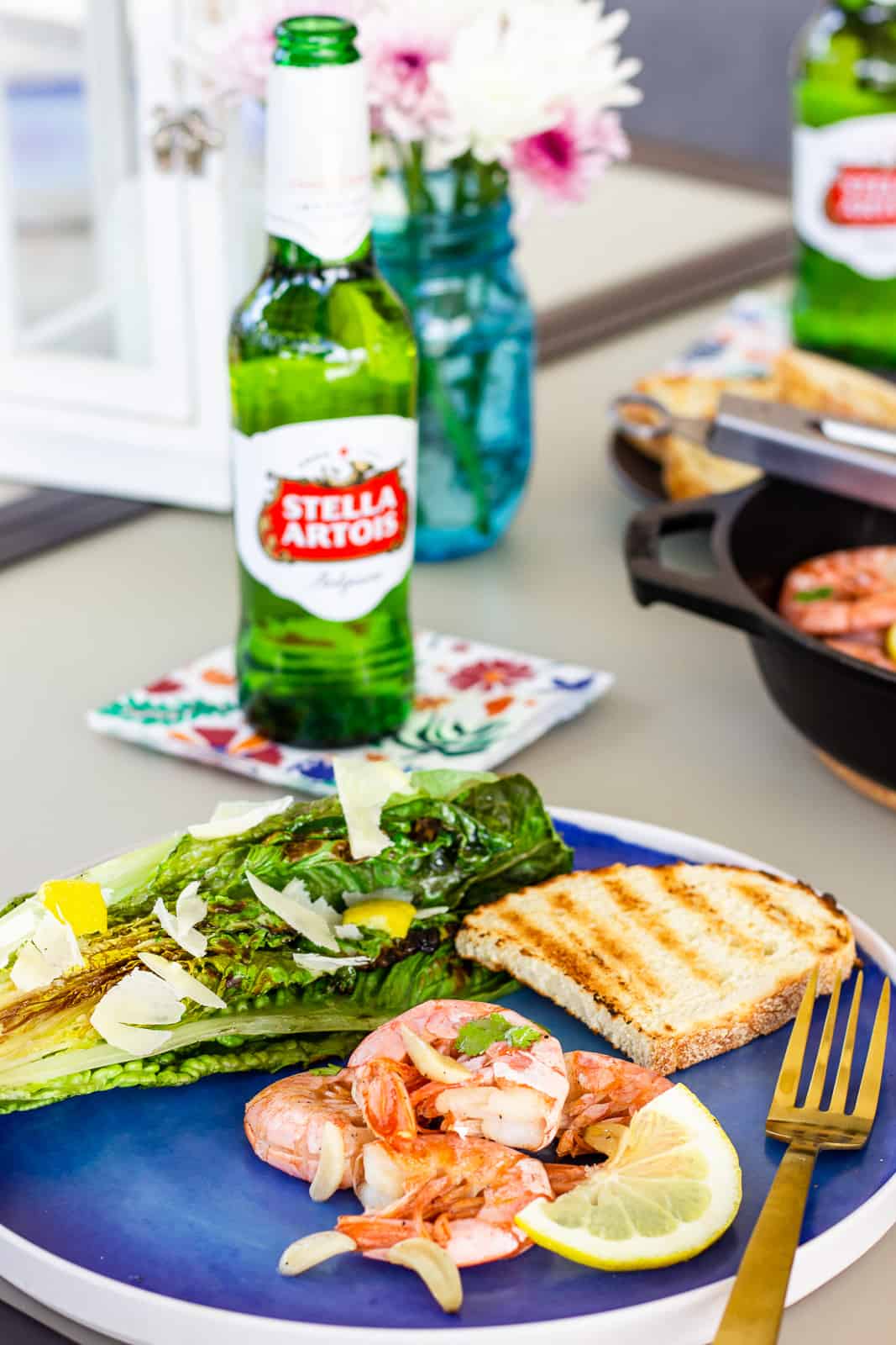 Blue plate with shrimp, slice of grilled bread, halved grilled romaine, and a bottle of Stella Artois Beer.