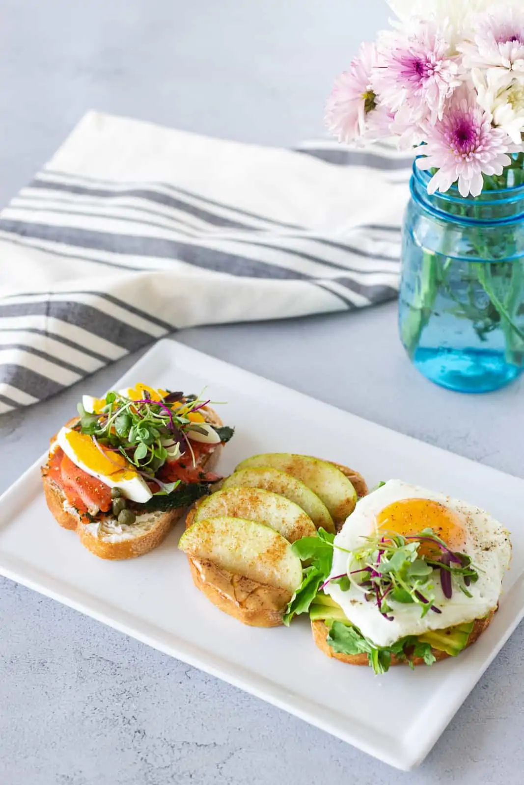 White plate with 3 different toasts, lox, peanut butter apple, and avocado toast. Blue mason jar with flowers on the side.