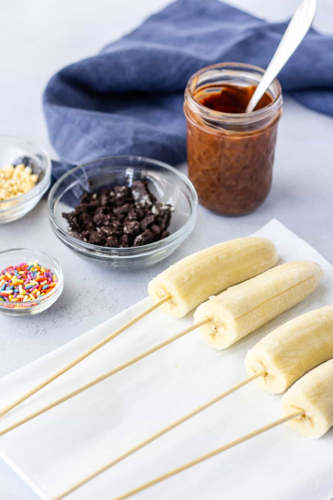 Halved bananas on skewers on a plate with a jar of melted chocolate and toppings in bowls.