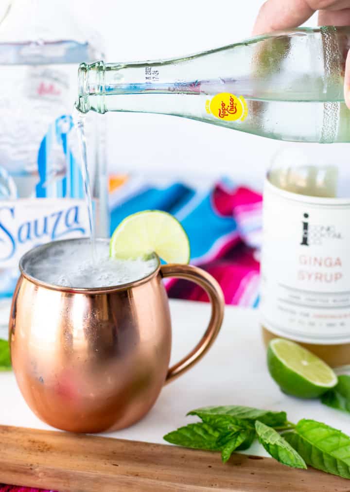 Hand pouring a glass bottle of topo chico into a copper mug.