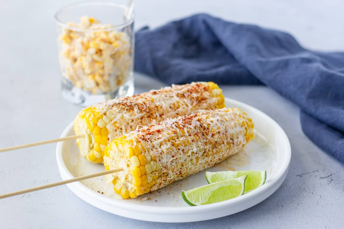 2 ears of corn with lime wedges on the plate and a glass of shaved corn.