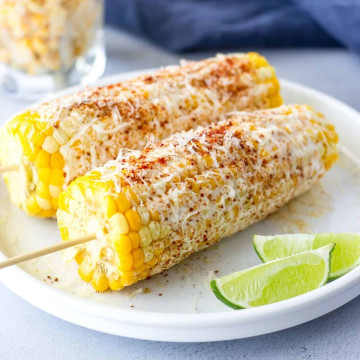 2 Ears of corn with skewers on a plate with lime wedges on the side.