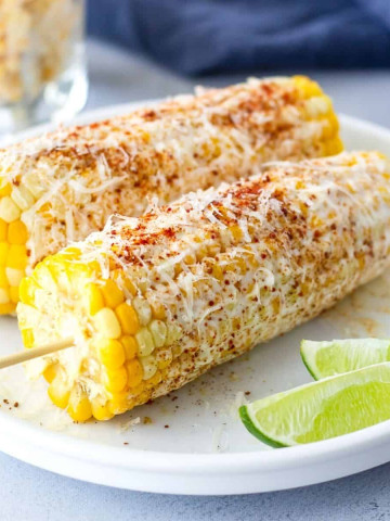 2 Ears of corn with skewers on a plate with lime wedges on the side.
